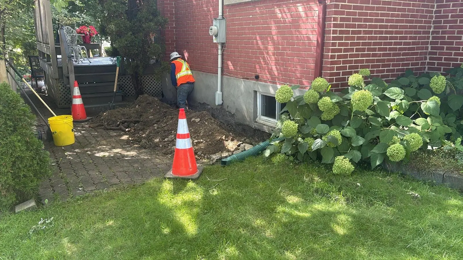 Excavation along a brick house foundation, with safety cones and a worker at the trench