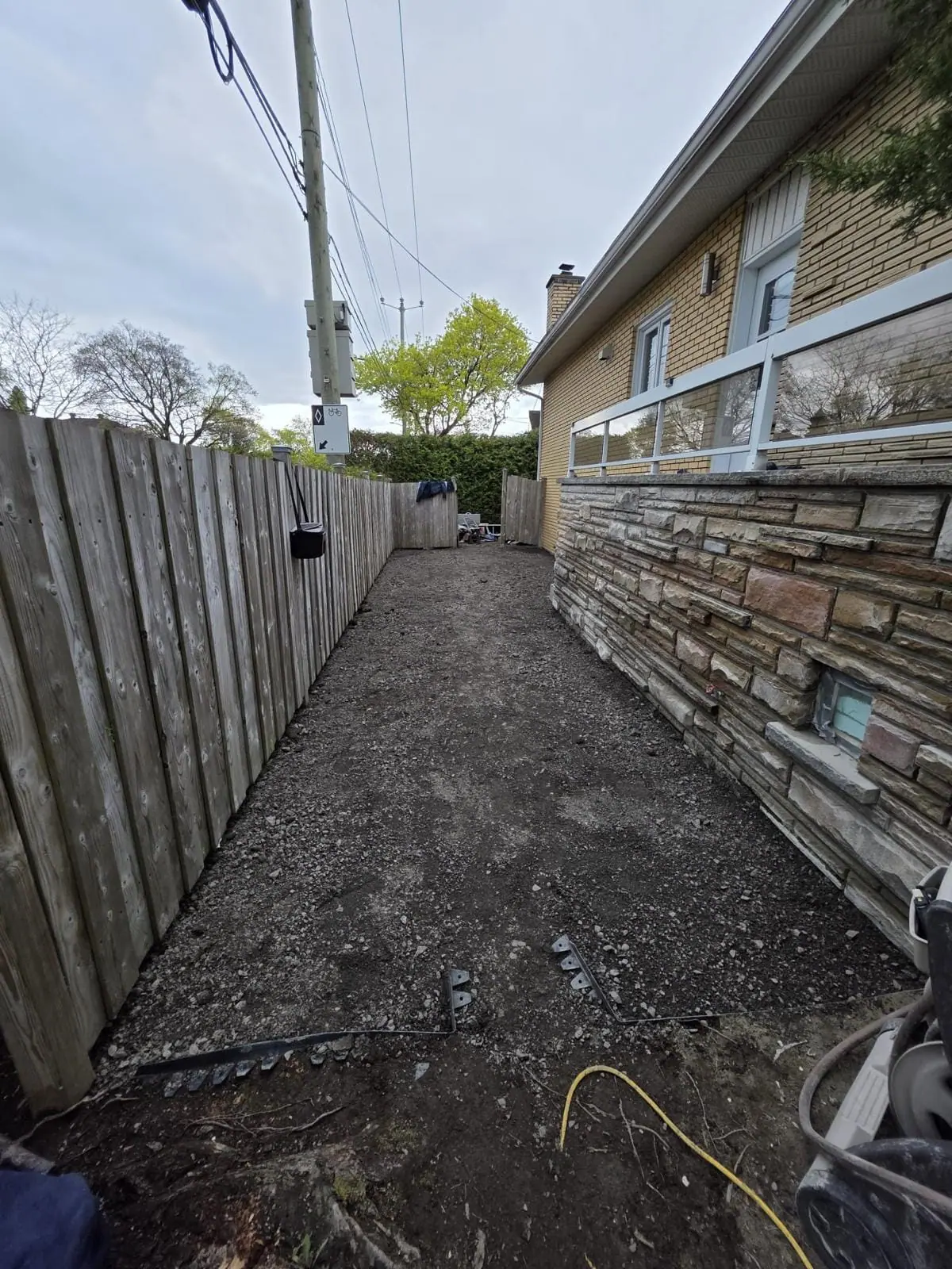 Side yard before paving: compacted gravel base beside a stone-clad wall and fence